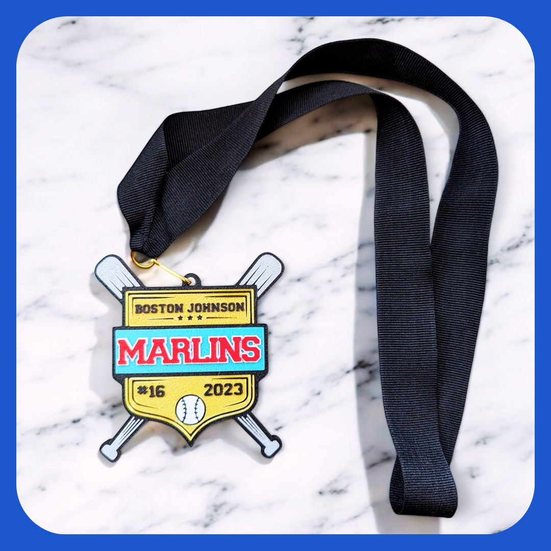 A custom baseball medal with a black ribbon, featuring a shield-shaped design with crossed baseball bats in the background. The medal is gold with turquoise and red accents, displaying the name 'Boston Johnson,' the team name 'Marlins,' the number '#16,' and the year '2023,' along with a small baseball icon at the bottom. The medal is placed on a marble surface.